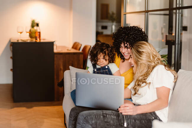 Excited young lesbian couple and cute child watching funny video on netbook and sitting on comfy couch in living room — Stock Photo
