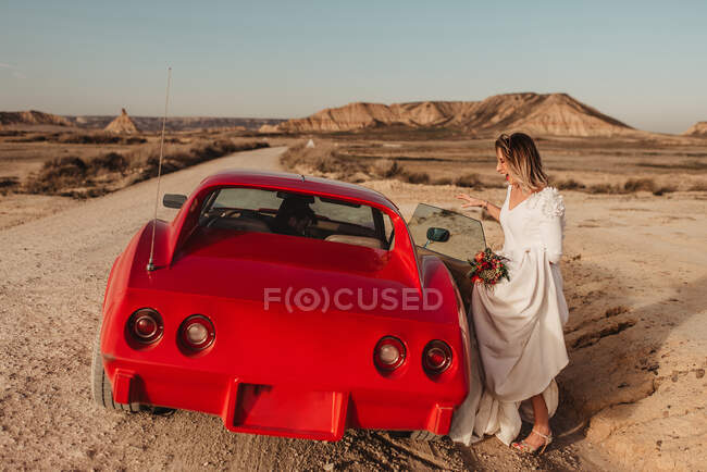 Donna felice corpo intero in abito bianco entrare nel lussuoso veicolo rosso parcheggiato su strada polverosa durante il viaggio attraverso il Parco Naturale Bardenas Reales in Navarra, Spagna — Foto stock