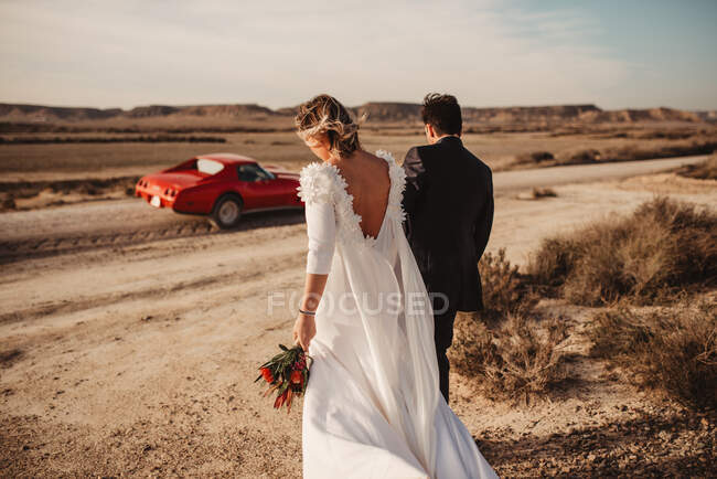 Vista posteriore di sposi anonimi che passeggiano insieme verso un'auto rossa di lusso nel deserto del Parco Naturale di Bardenas Reales in Navarra, Spagna — Foto stock