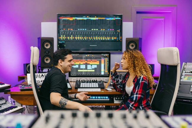 Side view of sound man talking to cheerful ethnic female partner while looking at each other among professional equipment in recording studio — Stock Photo