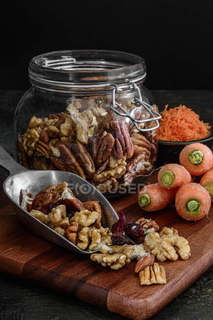 Tarro de vidrio con nueces y frutas secas sobre un fondo rústico oscuro junto a las zanahorias, ingredientes para preparar un pastel de zanahoria - foto de stock