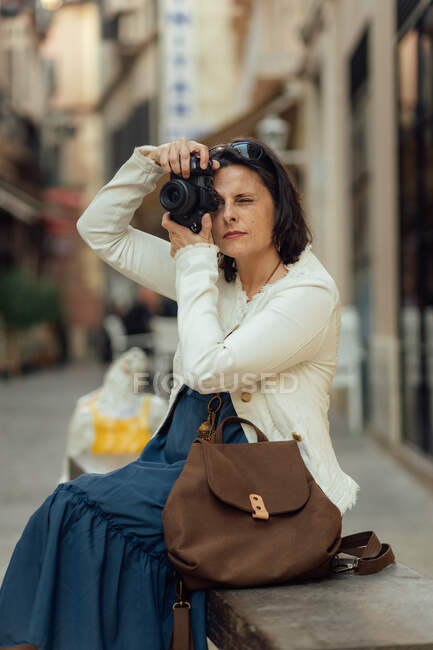 Viajero femenino adulto en ropa elegante con bolso que toma fotos en la cámara fotográfica mientras que está sentado en el banco contra fondo urbano borroso - foto de stock