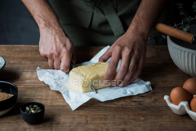 Unrecognizable man using knife to cut piece of butter while preparing pastry on wooden table near eggs and seeds — Stock Photo