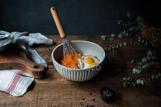 Batir el utensilio en un tazón con puré de calabaza, huevos y harina para la preparación del pastel en la mesa de madera cerca de la tabla de cortar y la toalla - foto de stock