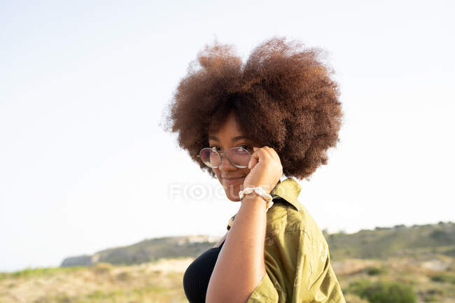 Vista laterale di giovane donna afroamericana sorridente con i capelli ricci che toccano gli occhiali e guardando la fotocamera mentre si gode le vacanze estive in campagna — Foto stock