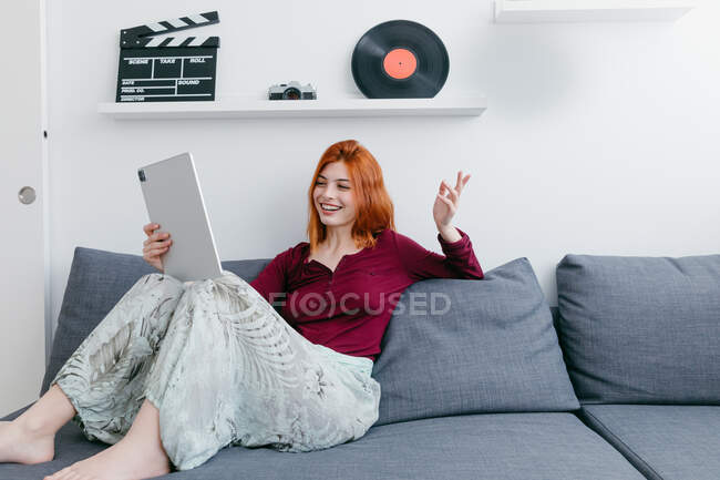 Contenido joven mujer sentada en el sofá mientras habla con su pareja durante el chat de vídeo en la tableta en casa - foto de stock