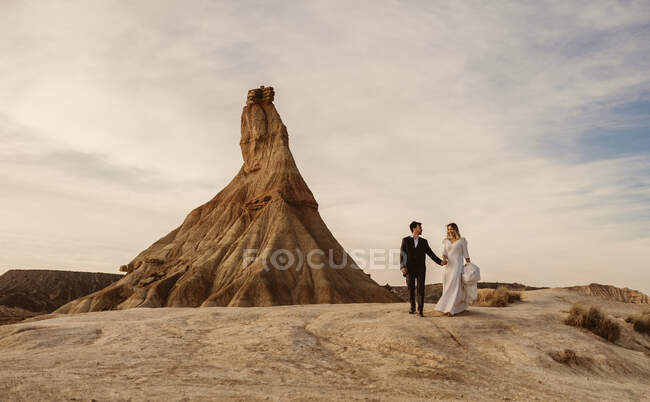 Happy groom and bride walking holding hands near mountain against cloudy sundown sky in Bardenas Reales Natural Park in Navarra, Spain — Stock Photo