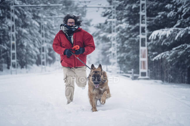 Hombre en chaqueta de esquí corriendo con perro doméstico con plomo en la nieve entre los árboles en el bosque de invierno - foto de stock