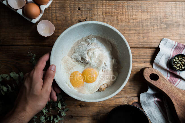 From above chef putting bowl with eggs and cream mixed with bread crumbs and flour on wooden table during pastry preparation — Stock Photo
