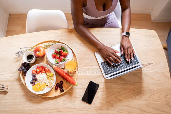Desde arriba de la cosecha anónima afroamericana escribiendo en netbook mientras está sentado en la mesa con nutritivo desayuno saludable en casa - foto de stock