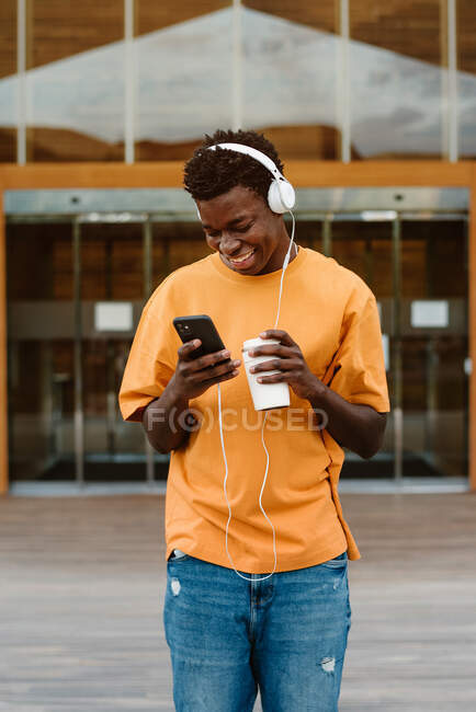 Happy African American male browsing on smartphone while listening to music in headphones walking against modern building — Stock Photo