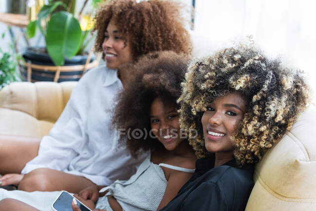 Счастливые афроамериканские женщины и девушка улыбаются, сидя на диване в выходные вместе — стоковое фото