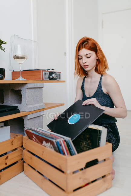 Junge Frau wählt Schallplatte aus Holzcontainer aus, während sie im Hauszimmer gegen Vintage-Plattenspieler sitzt — Stockfoto