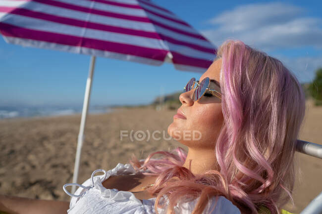 Вид сбоку расслабленной женщины с розовыми волосами, лежащей в шезлонге и загорающей во время летнего отпуска — стоковое фото