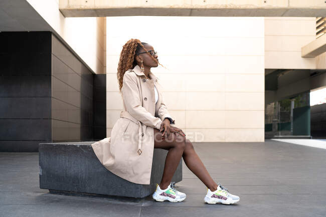 Side view full body of African American female with curly hair dressed in cloak and sneakers sitting near building and looking away — Stock Photo