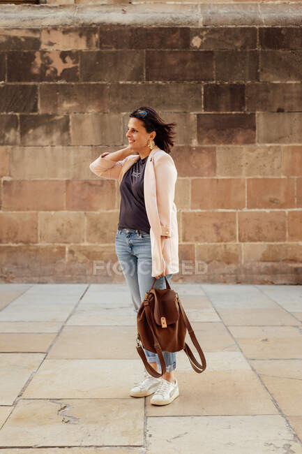 Full body of positive adult female in trendy casual outfit with backpack in hand standing against aged stone wall on urban street — Stock Photo