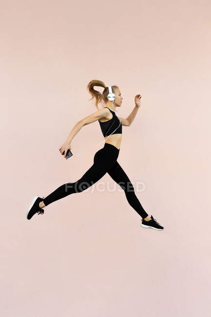 Joven atlética mujer caucásica con auriculares y ropa deportiva, saltando sobre un fondo brillante al aire libre - foto de stock