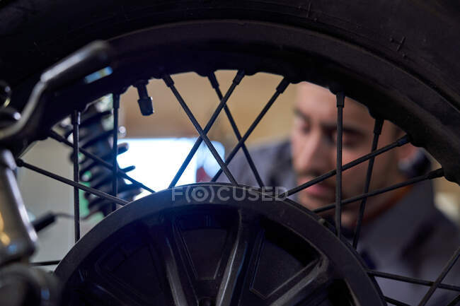 Trabajador varón adulto con mono que fija la rueda trasera de la motocicleta mientras está parado en el banco de trabajo en el taller - foto de stock