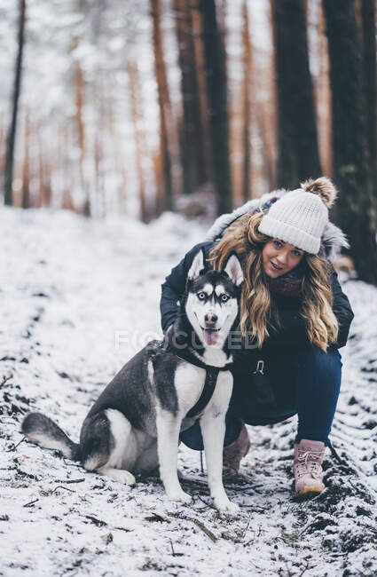 Frau in Skijacke geht mit Hund im Winterwald zwischen Bäumen spazieren — Stockfoto
