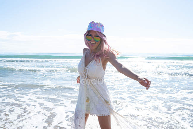 Conteúdo fêmea com cabelo rosa e vestido de verão desfrutando de liberdade com braços estendidos enquanto está de pé na praia contra o mar e olhando para a câmera — Fotografia de Stock
