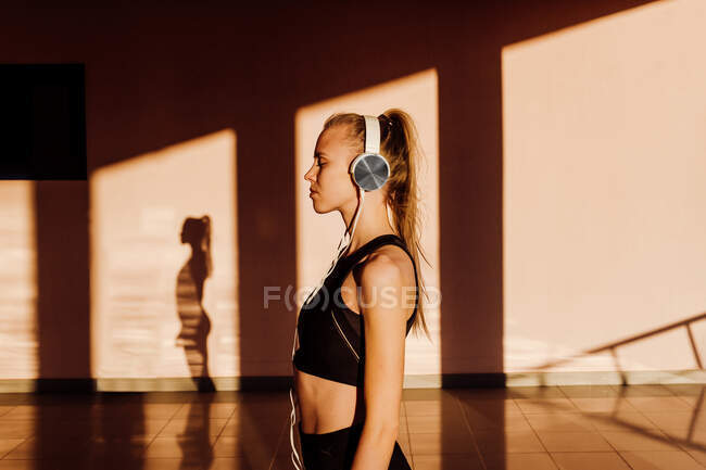 Joven atlética mujer caucásica escuchando música con los ojos cerrados y relajándose al atardecer, las sombras y la luz en el fondo - foto de stock