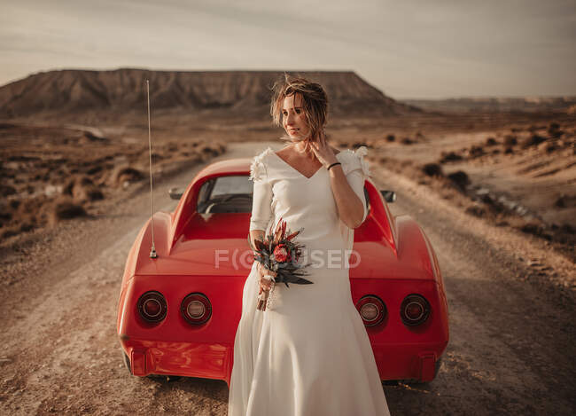 Счастливая женщина в белом платье, стоящая в стороне от роскошного красного автомобиля, припаркованного на пыльной дороге во время поездки через природный парк Барденас-Реалес в Наварре, Испания — стоковое фото