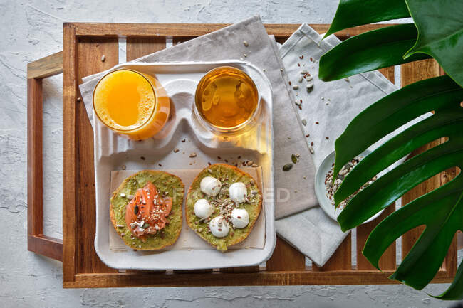 Сверху вкусные тосты из авокадо с лососиной и сыром буррата, подаваемые на столе со стаканами свежего сока и травяного чая, размещенного рядом с экзотическим растением Monstera deliciosa — стоковое фото