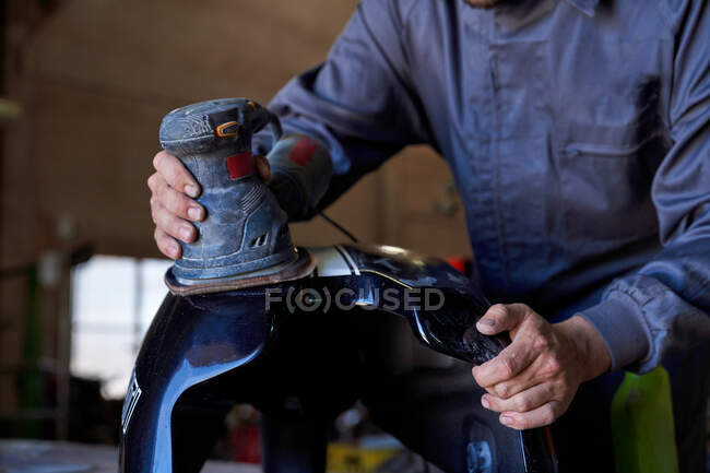 Нерозпізнані чоловіки стоять на робочому місці і полірують мотоциклетний танк за допомогою електричного інструмента під час роботи в гаражі. — стокове фото
