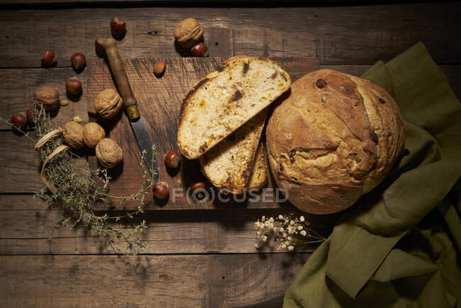 Верхний вид вкусного домашнего хлеба из теста с орехами, помещенными на деревянную доску с ножом и ароматическими травами на деревенском столе — стоковое фото