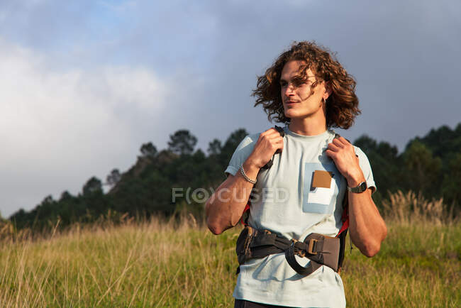 Mochilero masculino caminando en el prado al atardecer durante el trekking en verano y mirando hacia otro lado - foto de stock