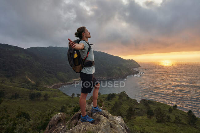 Vista lateral del caminante masculino despreocupado de pie en el mirador sobre la roca y disfrutando de la libertad en el fondo de la puesta de sol sobre el mar - foto de stock