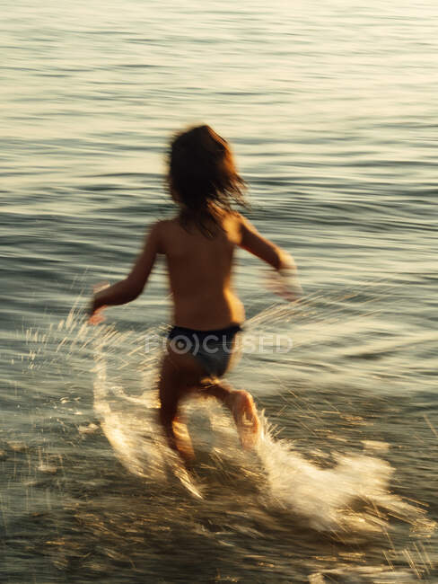Visão traseira da criança irreconhecível em trajes de banho correndo para a água do mar com salpicos enquanto desfruta de verão na praia — Fotografia de Stock