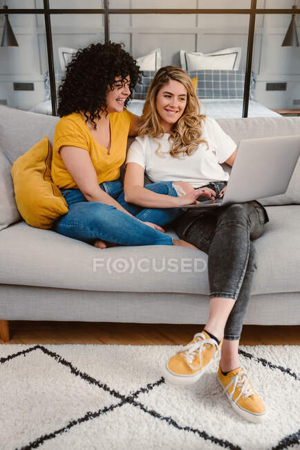 Cuerpo completo riendo joven lesbiana pareja navegación netbook juntos mientras sentado en acogedor sofá en sala de estar - foto de stock