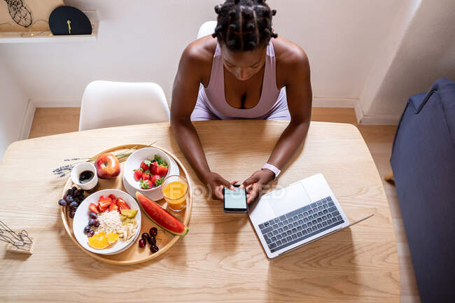 Von oben surft eine Afroamerikanerin am Handy, während sie zu Hause am Tisch mit reifen, gesunden Früchten sitzt — Stockfoto