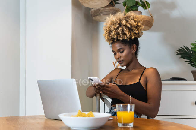 Atractiva mujer afroamericana freelancer sentada en la silla y viendo el ordenador portátil mientras navega en el teléfono inteligente en la mesa con desayuno - foto de stock