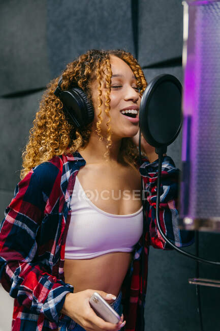 Contenu jeune chanteuse noire dans les écouteurs toucher le ventre tout en chantant dans le microphone dans le studio de musique — Photo de stock