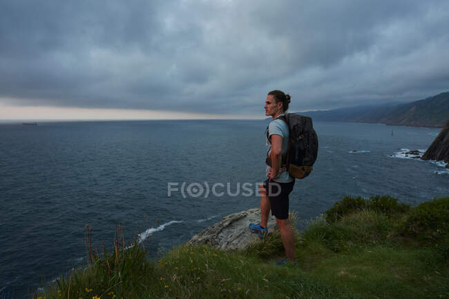 Vista lateral do viajante masculino em pé na rocha e admirando a vista do mar durante o trekking no verão — Fotografia de Stock