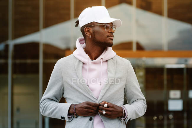 Confident African American male wearing casual clothes and cap and sunglasses standing against modern building and looking away — Stock Photo