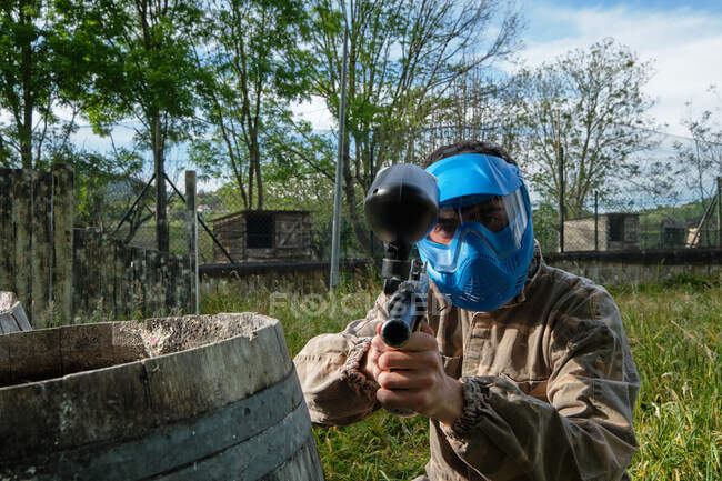 Male player in helmet and camouflage outfit aiming at camera during paintball game in countryside — Stock Photo