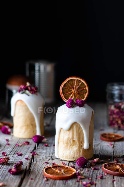 Несколько вкусных домашних кулихов, залитых сладкой глазурью и украшенных кусочками сухого апельсина и цветов на деревянном столе — стоковое фото