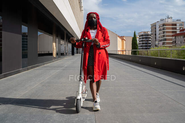 Cuerpo completo de mujer afroamericana en máscara con trenzas brillantes montando scooter eléctrico a lo largo del edificio de la torre en tiempo libre - foto de stock