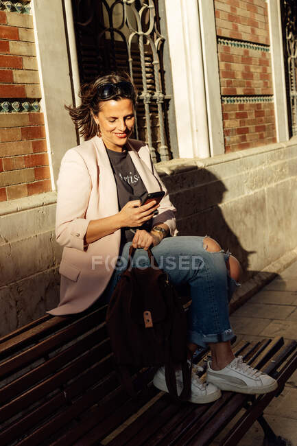 Mujer étnica adulta de moda positiva en jeans rasgados y chaqueta casual sentada en el banco cerca del edificio urbano y navegando por el teléfono móvil - foto de stock