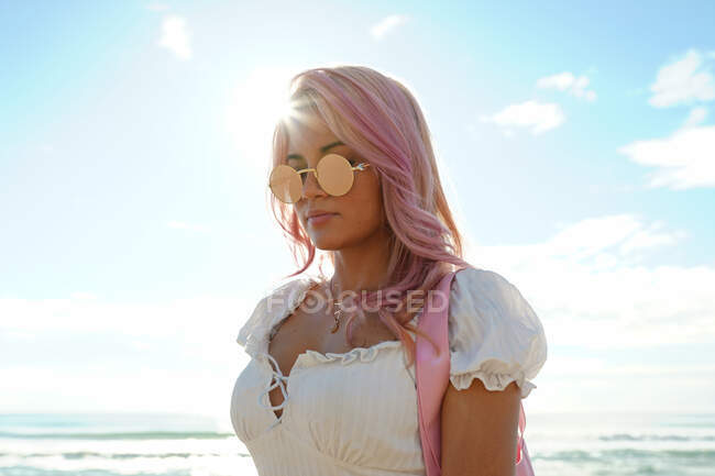 Donna alla moda con i capelli rosa e in occhiali da sole rotondi in piedi su sfondo di mare e cielo blu durante le vacanze estive — Foto stock