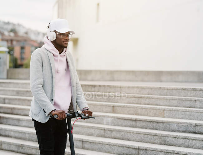 Focus thoughtful African American male in wireless headphones riding electric scooter near stairway in city — Stock Photo
