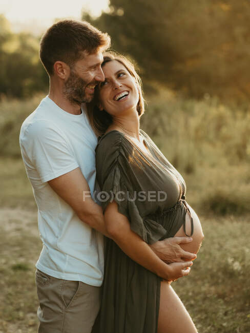 Side view of smiling male hugging pregnant female from behind while standing in countryside meadow at sunset — Stock Photo