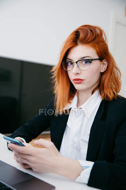 Junge Unternehmerin in formeller Kleidung trägt SMS auf dem Handy, während sie zu Hause am Schreibtisch mit Laptop arbeitet — Stockfoto