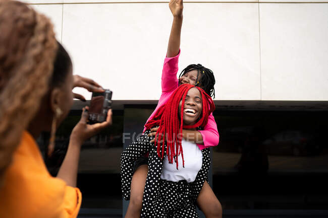 Low angle of crop African American female taking picture on photo camera of laughing girlfriend giving piggyback to best friend with hand raised — Stock Photo