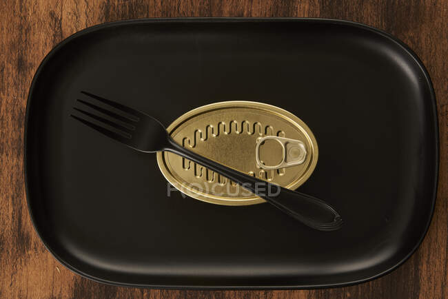Верхний вид черной вилки, расположенной рядом с запечатанной консервной пищей на прямоугольном черном подносе — стоковое фото