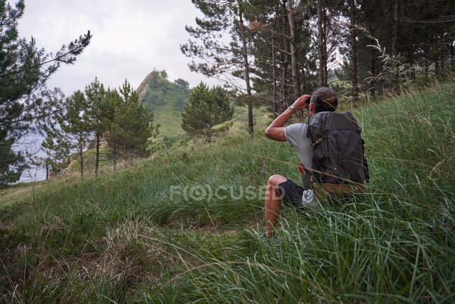 Back view of anonymous male backpacker sitting on hill and observing mountain top in binoculars during trekking in summer — Stock Photo