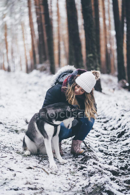 Dame en veste de ski marche avec chien domestique entre les arbres dans la forêt d'hiver — Photo de stock